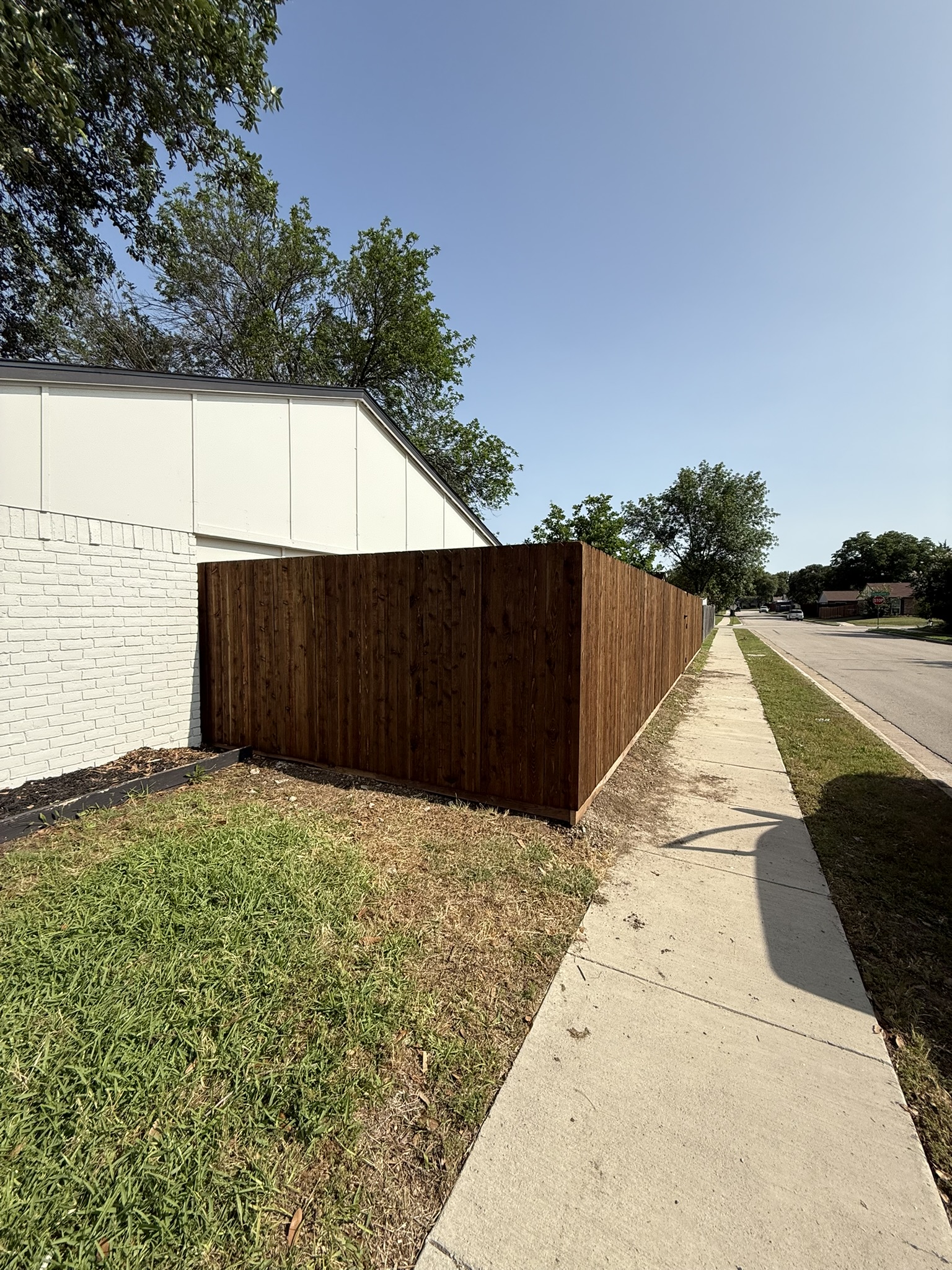 Dark-stained cedar board-on-board privacy fence along a sidewalk next to a white building