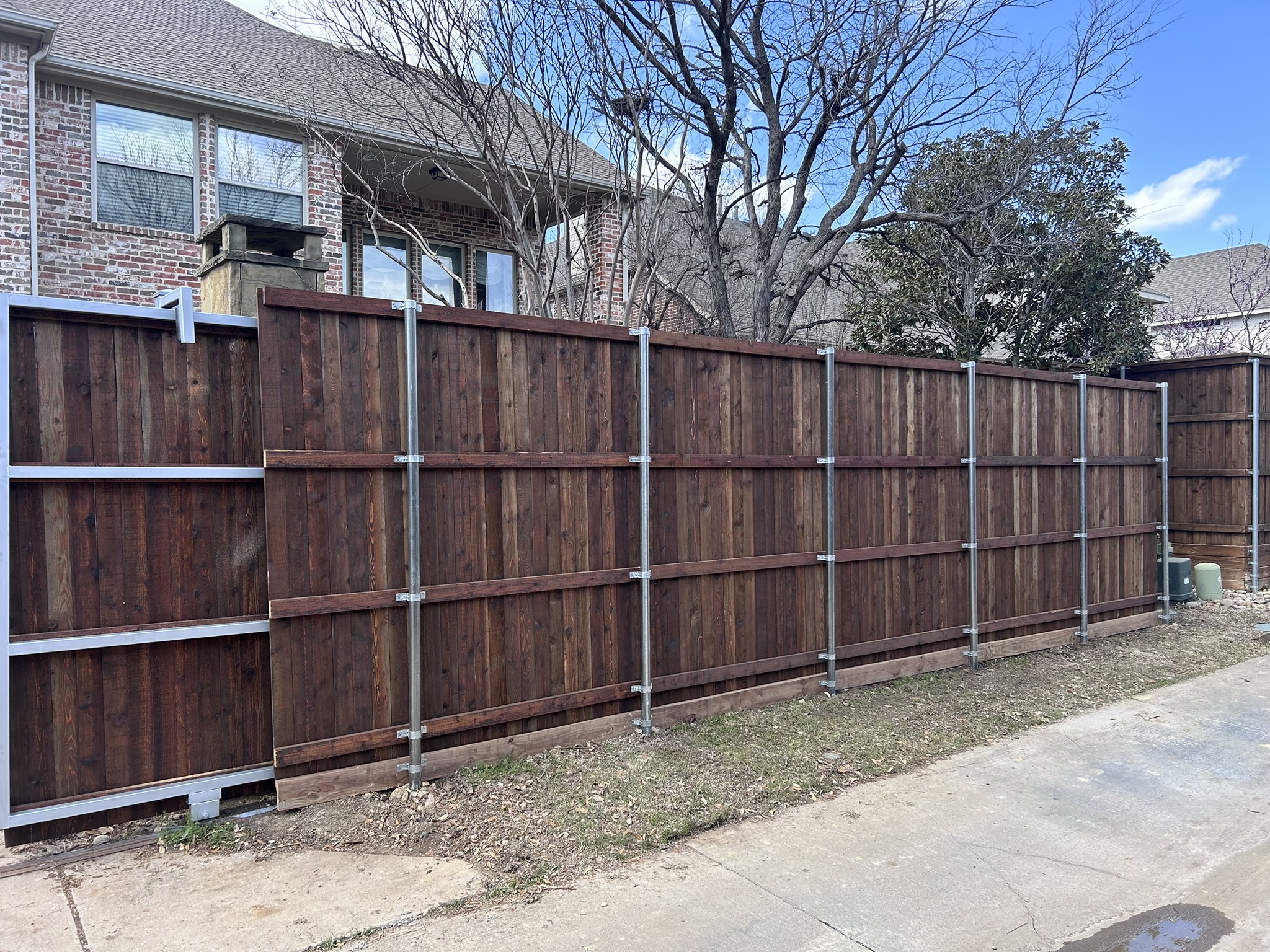 Back side of dark-stained cedar fence showing metal post framework along a sidewalk