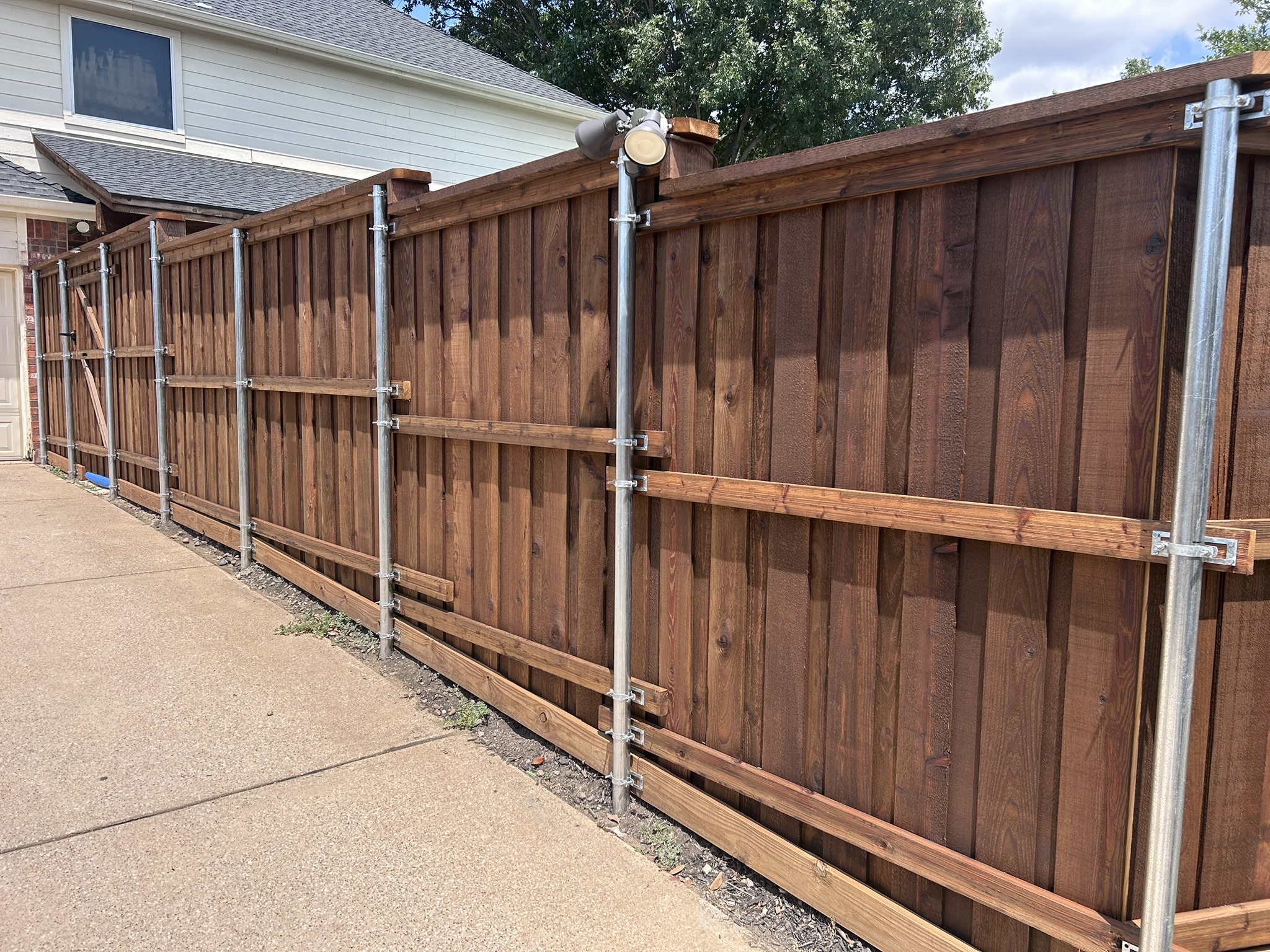 Back side of stained cedar board-on-board fence with metal posts along driveway